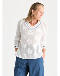 A woman wearing a white open-knit long-sleeve sweater with a blue and white V-neck trim, paired with blue pants, against a light gray studio background.