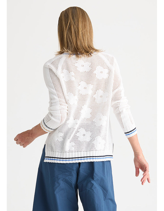 A woman shown from the back wearing a white open-knit cardigan with a floral pattern and blue pants, against a light gray studio background.