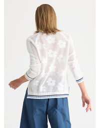 A woman shown from the back wearing a white open-knit cardigan with a floral pattern and blue pants, against a light gray studio background.