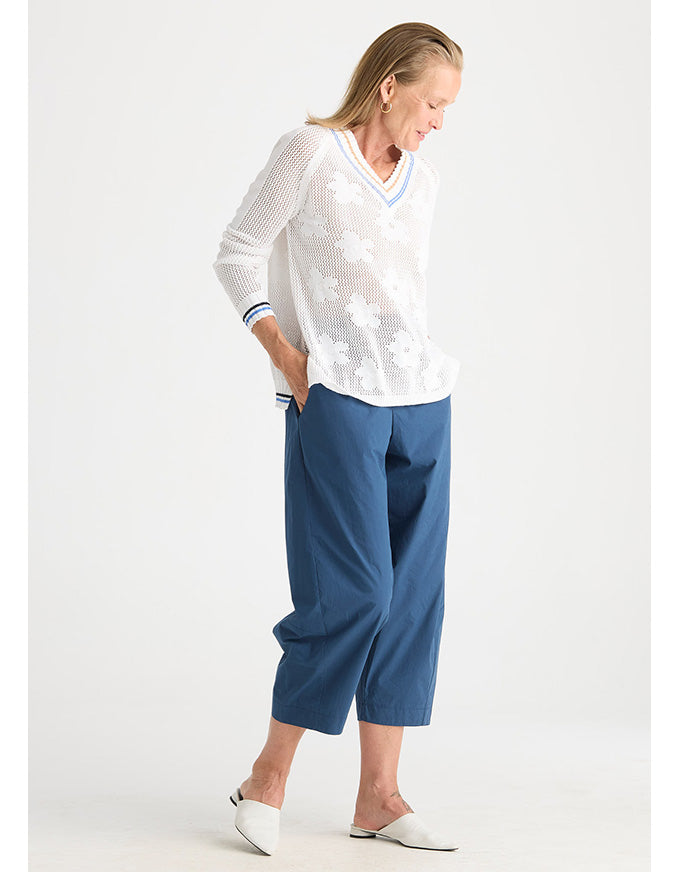 A woman modeling a white long-sleeve knit top with a V-neck and white floral embroidery, paired with blue cropped pants and white pointed flats, against a light gray studio background.