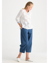 A woman modeling a white long-sleeve knit top with a V-neck and white floral embroidery, paired with blue cropped pants and white pointed flats, against a light gray studio background.