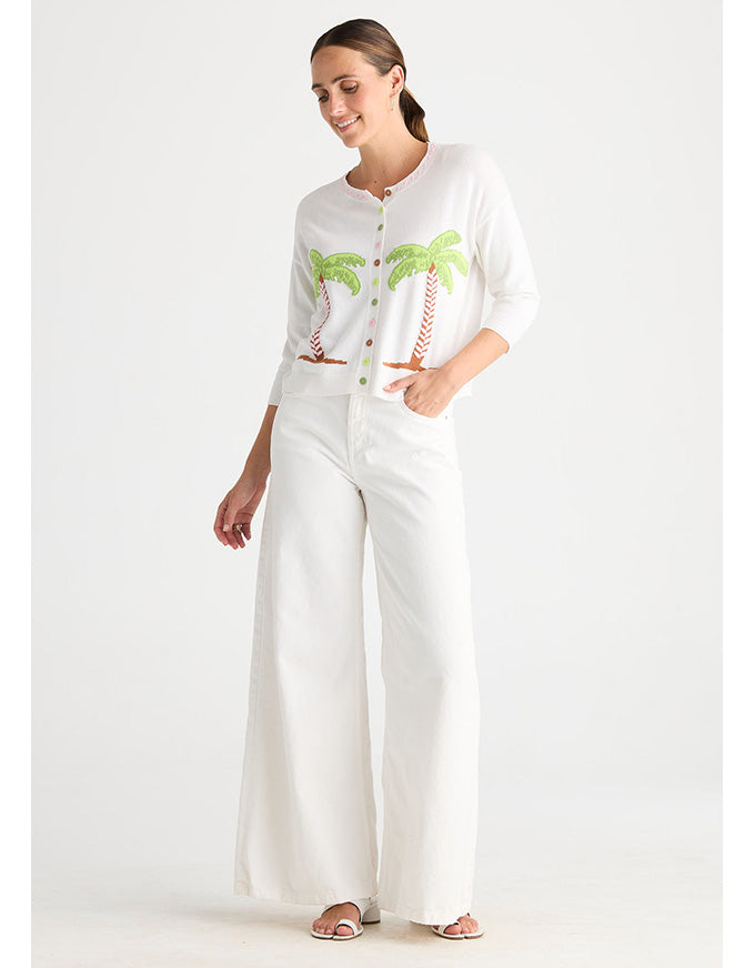 A woman modeling a white outfit consisting of a short-sleeve button-up cardigan with green palm tree embroidery, matching white wide-leg pants, and white heeled sandals, set against a plain white studio background.