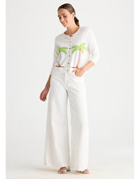 A woman modeling a white outfit consisting of a short-sleeve button-up cardigan with green palm tree embroidery, matching white wide-leg pants, and white heeled sandals, set against a plain white studio background.