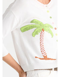 White knit cardigan sweater featuring a large green and orange palm tree motif on the front, with pastel-colored buttons along the neckline, shown on a model.