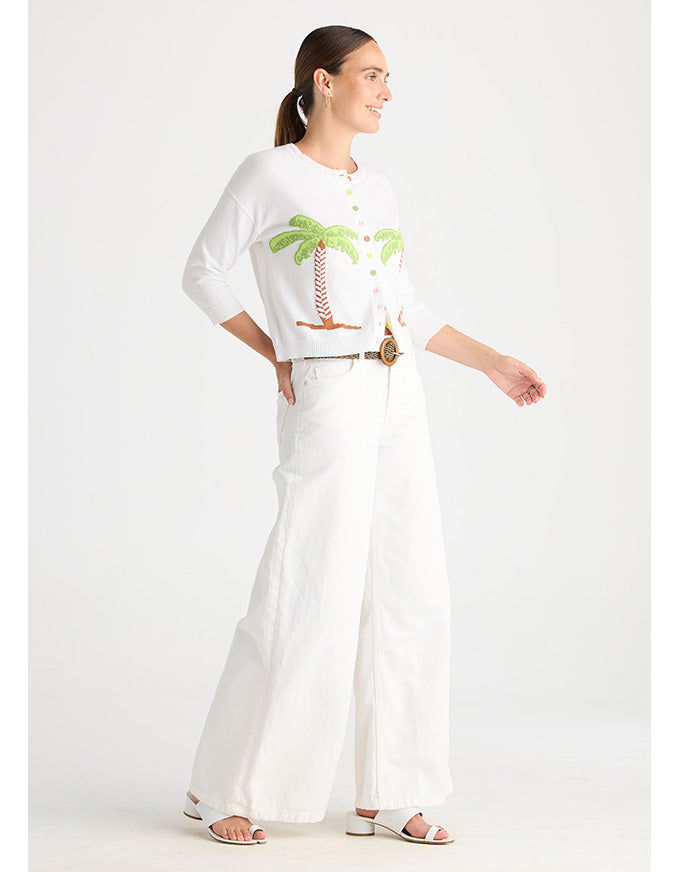 A woman modeling a white three-quarter sleeve top with a green palm tree graphic and white wide-leg pants, styled with a thin brown belt and white sandals against a light gray studio background.