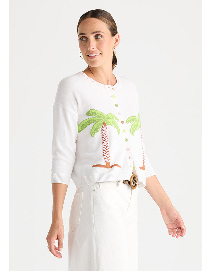 A woman modeling a white knit cardigan with green palm tree embroidery and white trousers against a light gray studio background.