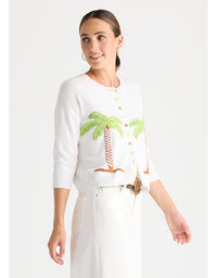 A woman modeling a white knit cardigan with green palm tree embroidery and white trousers against a light gray studio background.