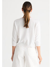 A woman viewed from the back wearing a white knit sweater with three-quarter sleeves and white pants, against a plain light gray studio background.