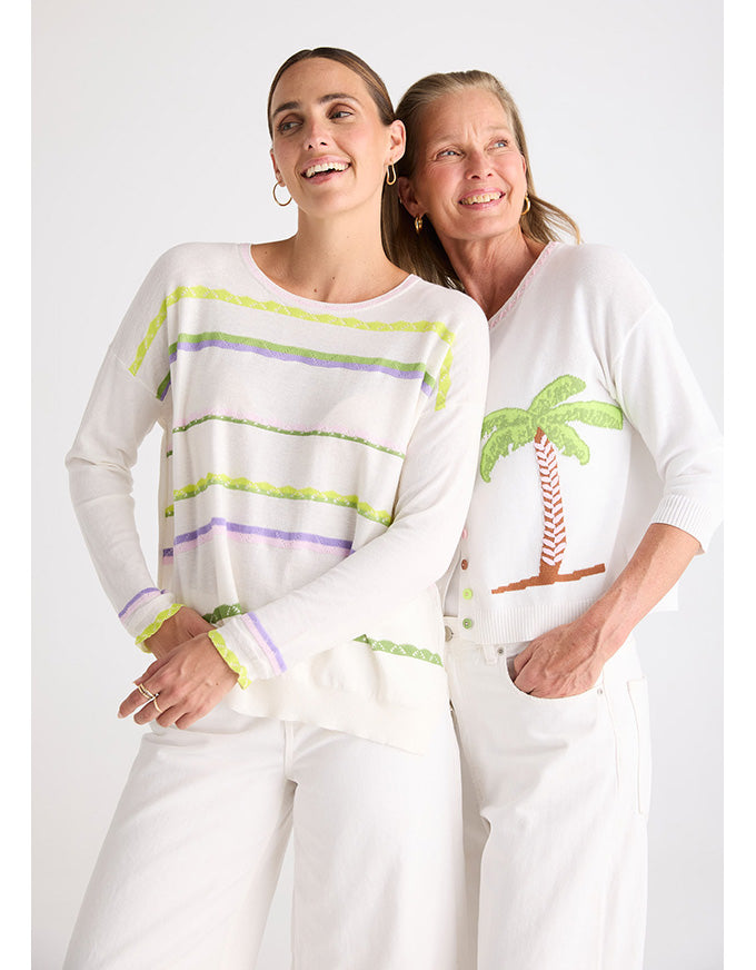 Two women modeling white knit sweaters with colorful stripe and palm tree graphics against a light gray studio background.
