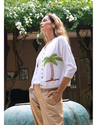 A woman wearing a white knit cardigan with a green palm tree motif and tan pants, standing outdoors under a leafy vine-covered structure.