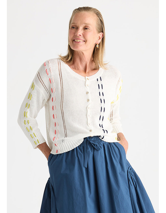 A woman wearing a white knit cardigan with colorful embroidered details and a blue skirt, photographed against a light gray studio background.