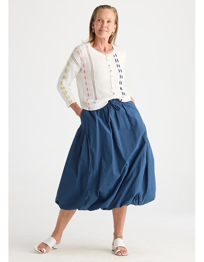 A woman modeling a white knit cardigan with colorful embroidered stripes and a blue midi skirt against a plain white studio background.