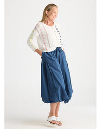 A woman modeling a white long-sleeve blouse with colorful embroidered details and high-waisted, voluminous blue harem-style pants against a plain light gray studio background.