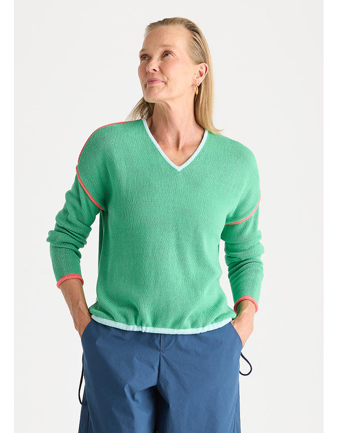 A woman wearing a green ribbed knit sweater with white and pink trim, paired with blue pants, against a light gray background.