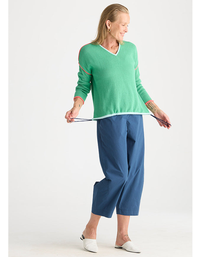 A woman modeling a green long-sleeve knit sweater with white trim and blue cropped pants against a plain light gray studio background.