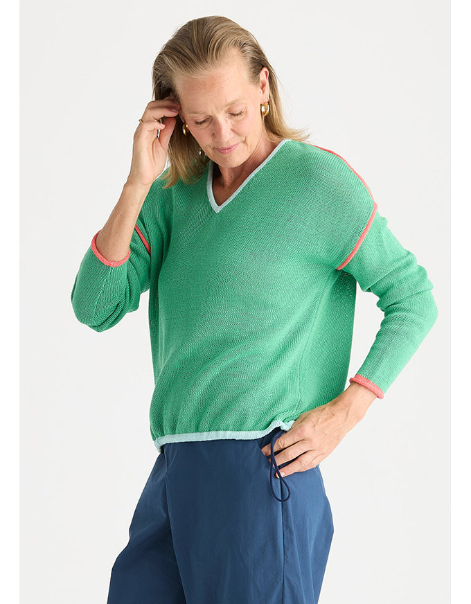 A woman wearing a green knit sweater with pink and white trim, paired with navy blue pants, photographed against a light gray studio background.