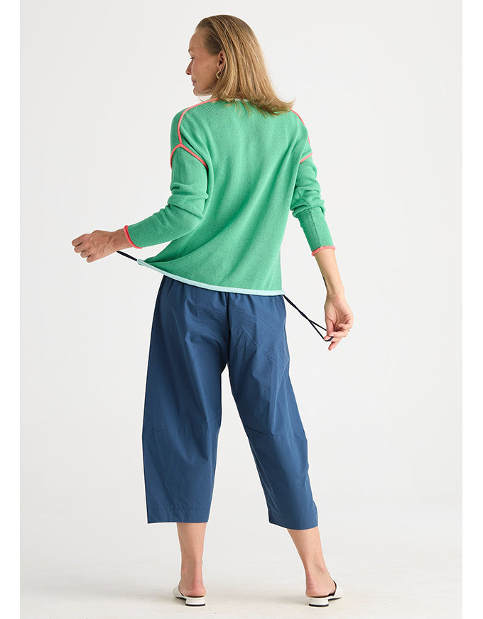 A woman shown from the back wearing a mint green knit cardigan with orange contrast stitching and blue cropped pants, standing against a light gray studio background.