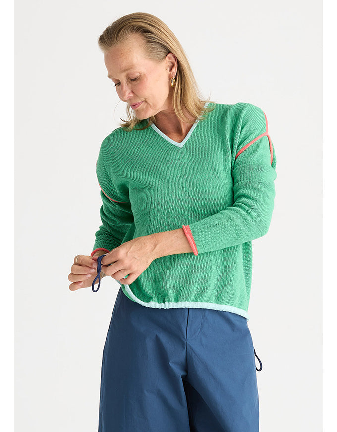 A woman wearing a green ribbed knit sweater with white and pink contrast trim, paired with blue pants, photographed against a light gray studio background.