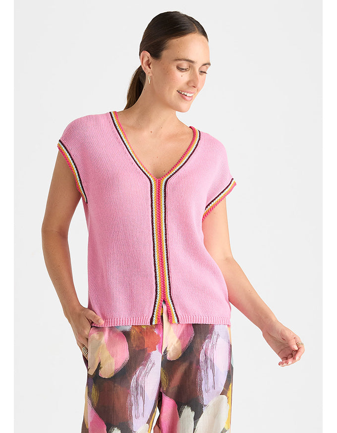 A woman wearing a pink ribbed knit short-sleeve top with colorful striped trim and a floral print skirt, photographed against a light gray studio background.
