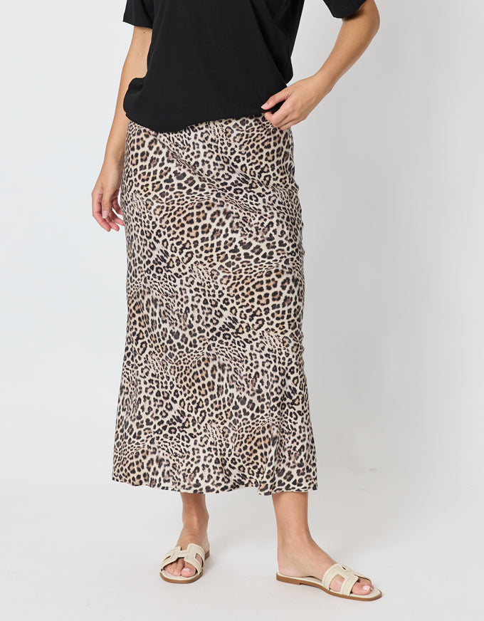 A model wearing a leopard print midi skirt with a black top and beige slide sandals, shown from the waist down against a light gray studio background.
