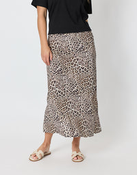 A model wearing a leopard print midi skirt with a black top and beige slide sandals, standing against a light gray studio background.
