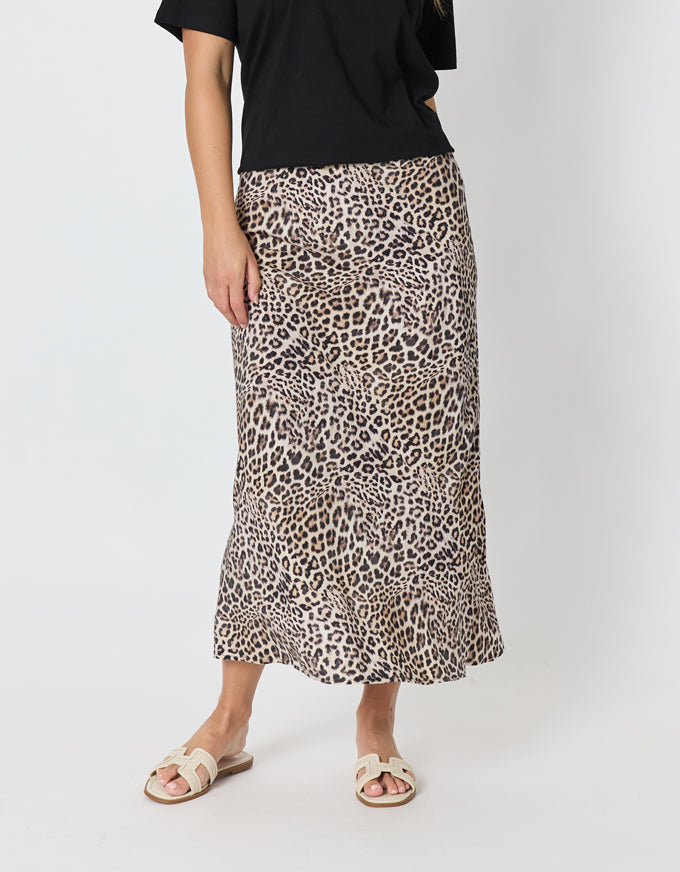 A model wearing a leopard print midi skirt with a black top and beige slide sandals, standing against a light gray studio background.