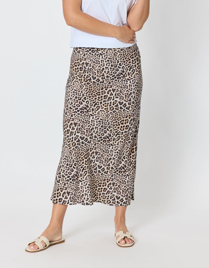 A model wearing a leopard print midi skirt with a white top and beige slide sandals, shown from the waist down against a light gray studio background.
