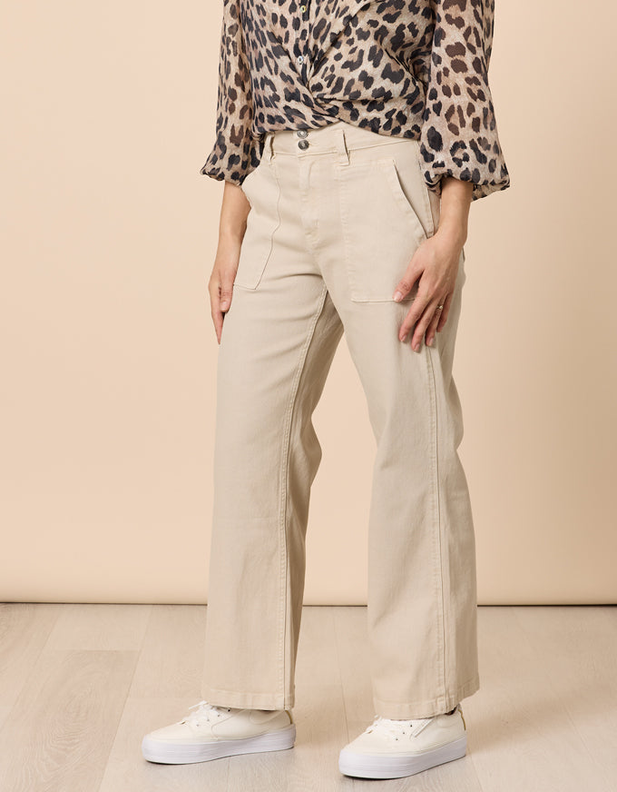 A person wearing beige wide-leg cropped pants, a leopard-print blouse, and white sneakers, standing against a plain beige studio wall and light wood floor.