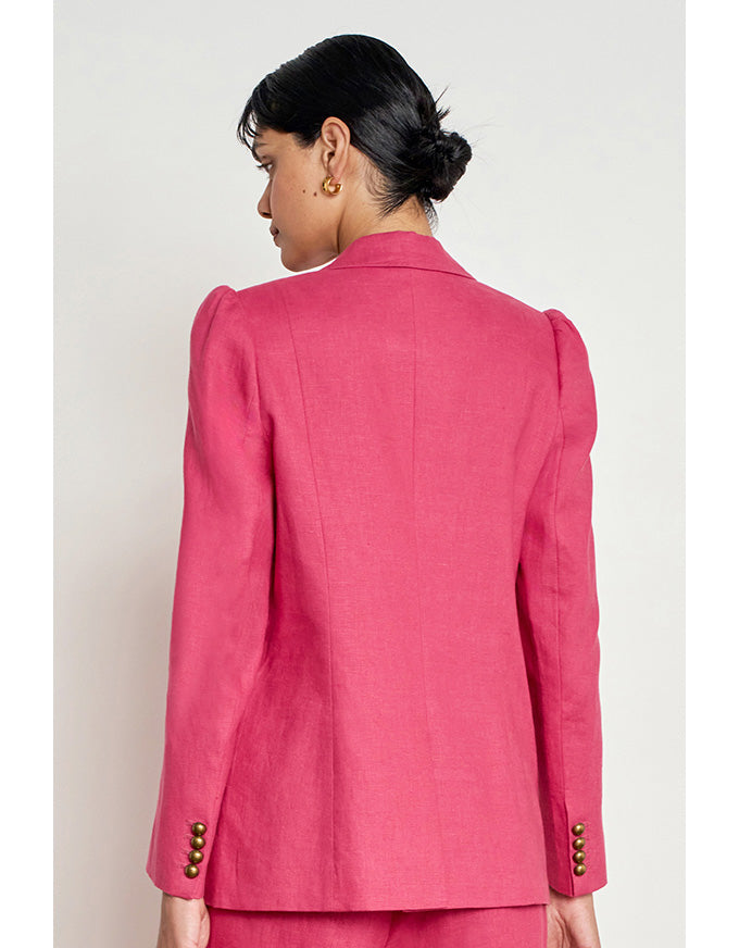 A woman shown from the back wearing a bright pink tailored blazer with structured shoulders and gold-tone button details at the cuffs, paired with matching pink bottoms against a plain light background. Crest Jacket Rhubarb