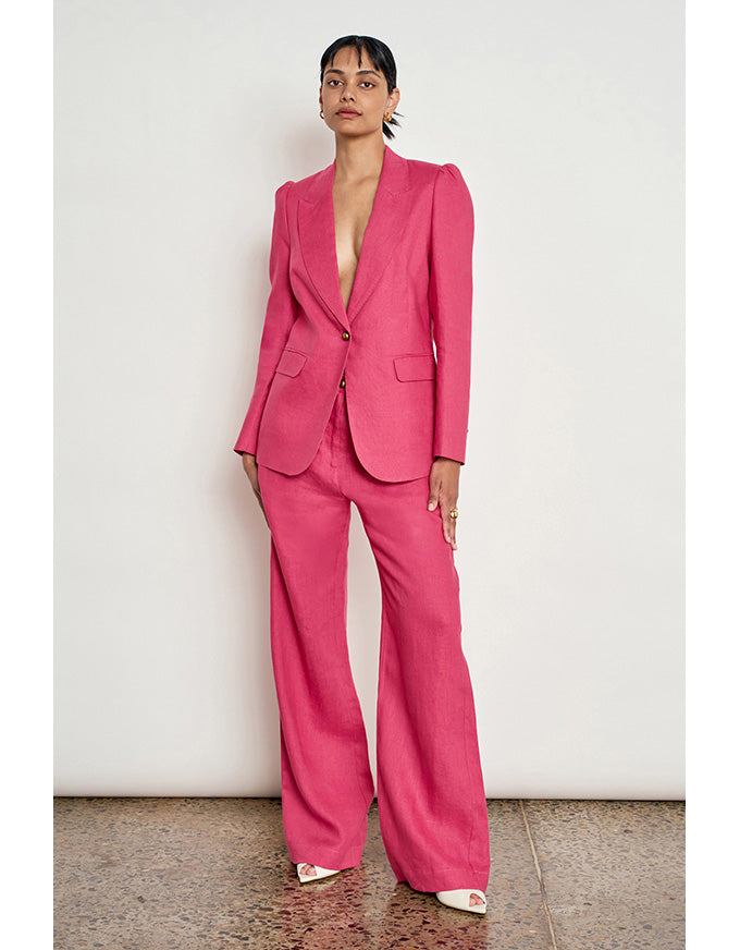 A model stands against a plain off-white wall wearing a bright pink tailored suit with a deep V-neck blazer and matching wide-leg trousers, paired with white pointed shoes. Crest Jacket Rhubarb