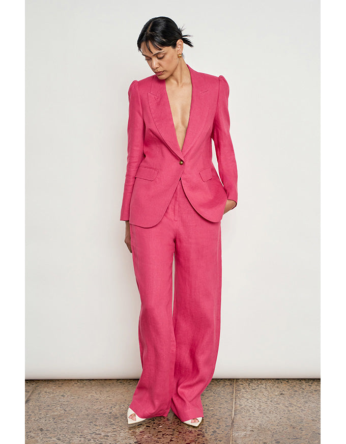 A model wearing a bright pink tailored suit with a deep V-neck blazer and matching wide-leg trousers, standing against a plain off-white wall. Edit Pant Rhubarb