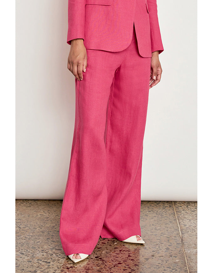 A model wearing a bright pink tailored pantsuit with wide-leg trousers and a matching blazer, paired with white pointed flats, standing against a plain off-white wall. Edit Pant Rhubarb
