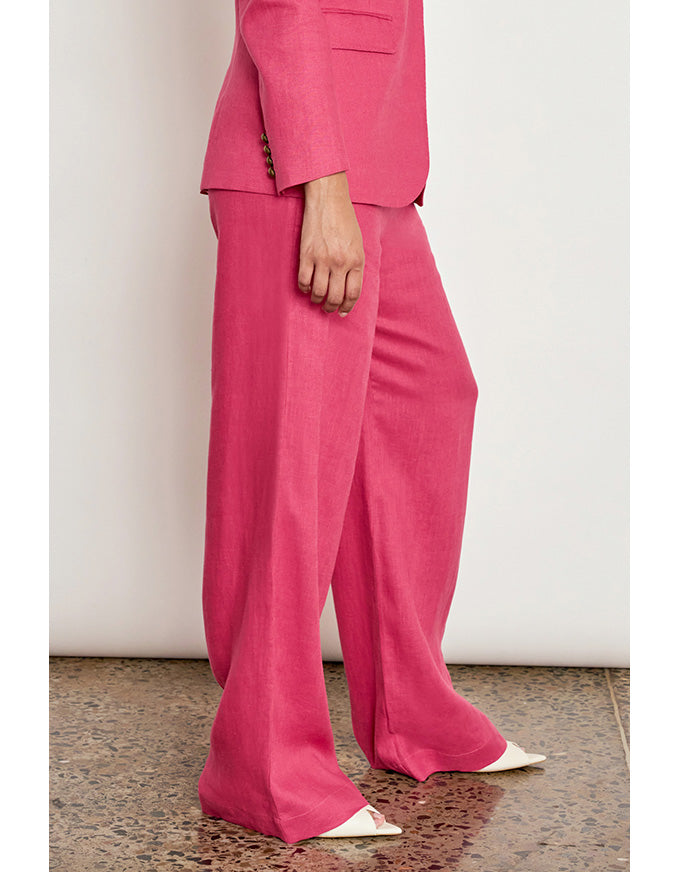 A model wearing a bright pink tailored suit with wide-leg trousers and white pointed shoes, standing against a plain off-white wall. Edit Pant Rhubarb