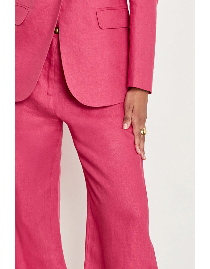 A close-up of a model wearing a bright pink tailored suit, showing the jacket lapels, a gold button, and the matching trousers, with one hand resting on the thigh. Edit Pant Rhubarb