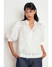 A woman wearing a white sheer embroidered blouse with puff sleeves and a split neckline, paired with dark blue jeans, standing against a plain off-white background. Poise Top Porcelain