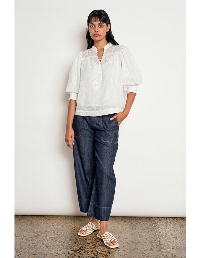 A woman stands against a plain off-white wall wearing a white embroidered blouse with puff sleeves, dark blue wide-leg cropped pants, and beige woven slide sandals. Poise Top Porcelain