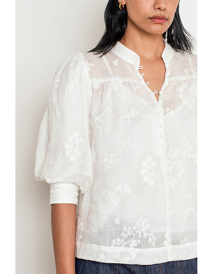 A woman wearing a white sheer embroidered blouse with puff sleeves and a split neckline, paired with dark blue jeans against a plain light background. Poise Top Porcelain