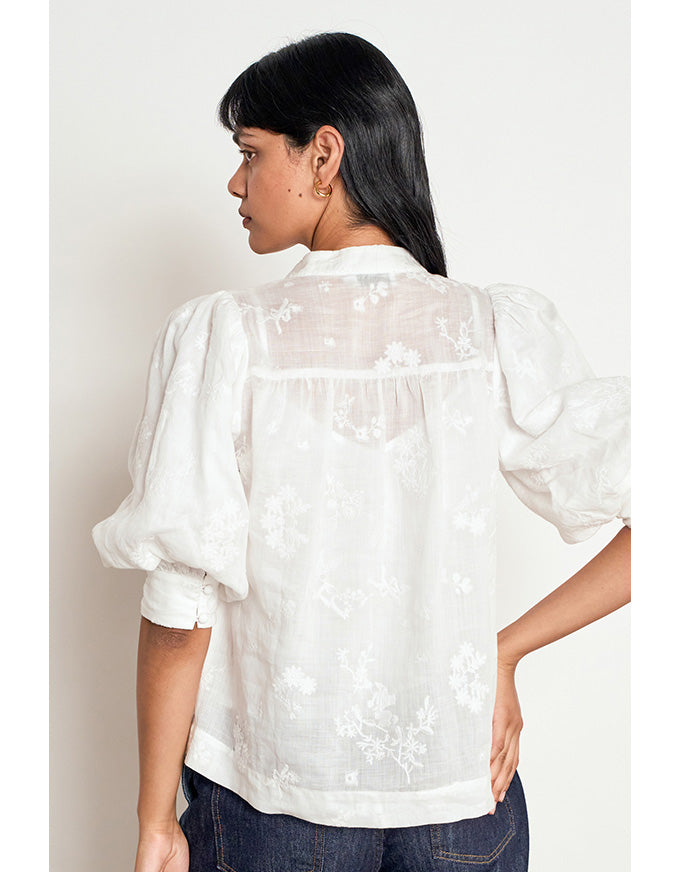A woman shown from the back wearing a white embroidered blouse with puff sleeves and a sheer yoke, paired with dark blue jeans against a plain light background. Poise Top Porcelain