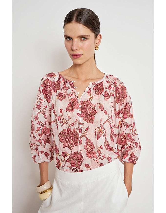 A woman wearing a light pink blouse with a red and burgundy floral pattern, paired with white trousers, photographed against a light gray studio background. Vow Top Rose Paisley