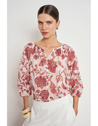 A woman wearing a light pink blouse with a red and burgundy floral pattern, paired with white trousers, photographed against a light gray studio background. Vow Top Rose Paisley
