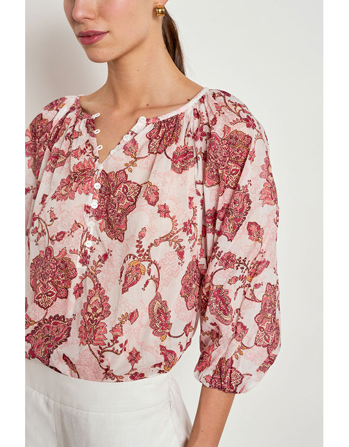 A close-up of a woman wearing a light pink blouse with a red and burgundy floral toile pattern, paired with white trousers. The blouse features a split neckline with decorative buttons and gathered puff sleeves. Vow Top Rose Paisley