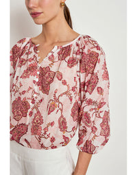 A close-up of a woman wearing a light pink blouse with a red and burgundy floral toile pattern, paired with white trousers. The blouse features a split neckline with decorative buttons and gathered puff sleeves. Vow Top Rose Paisley