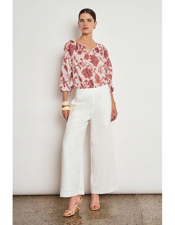 A woman stands against a plain off-white wall wearing a loose floral blouse with pink, red, and cream tones, paired with wide-leg white cropped pants and nude heeled sandals. Vow Top Rose Paisley