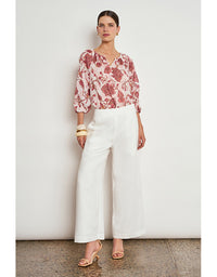 A woman stands against a plain off-white wall wearing a loose floral blouse with pink, red, and cream tones, paired with wide-leg white cropped pants and nude heeled sandals. Vow Top Rose Paisley