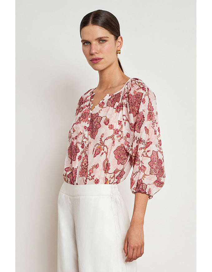 A woman modeling a light pink blouse with a red and burgundy floral pattern, paired with white trousers, against a plain light gray background. Vow Top Rose Paisley