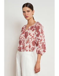 A woman modeling a light pink blouse with a red and burgundy floral pattern, paired with white trousers, against a plain light gray background. Vow Top Rose Paisley