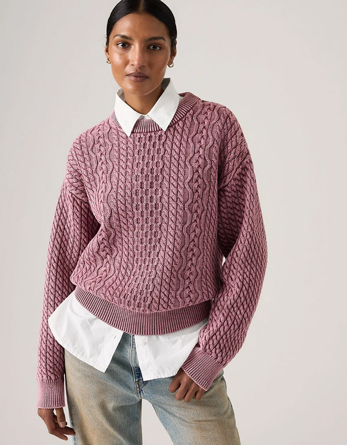 A woman wearing a dusty rose cable-knit cropped sweater over a white collared shirt and light-wash jeans, photographed against a plain light gray studio background.