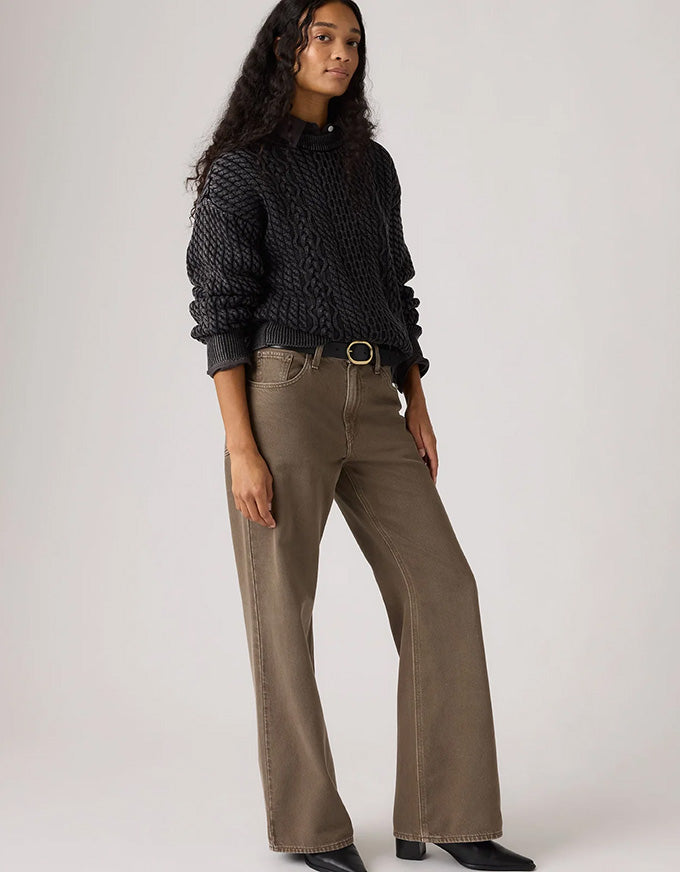 A model wearing a black textured knit mock-neck sweater and wide-leg brown pants with a gold-tone belt buckle, standing against a light gray studio background.