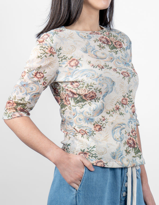 A woman wearing a beige three-quarter sleeve top with a blue and pink floral damask pattern, paired with light blue denim bottoms, against a plain light gray background.