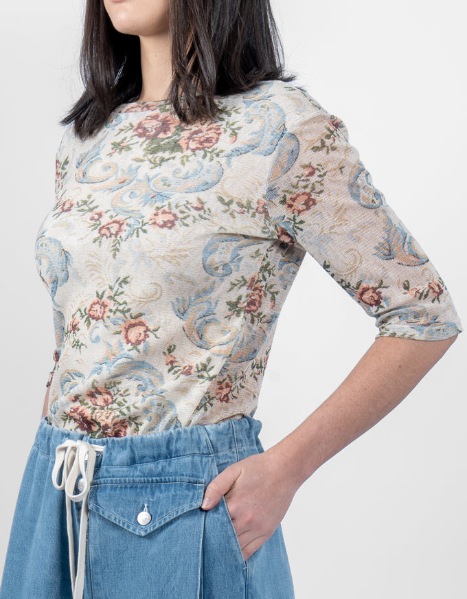 A woman wearing a beige short-sleeve top with a blue and pink floral paisley pattern, paired with blue denim shorts, against a plain light gray background.
